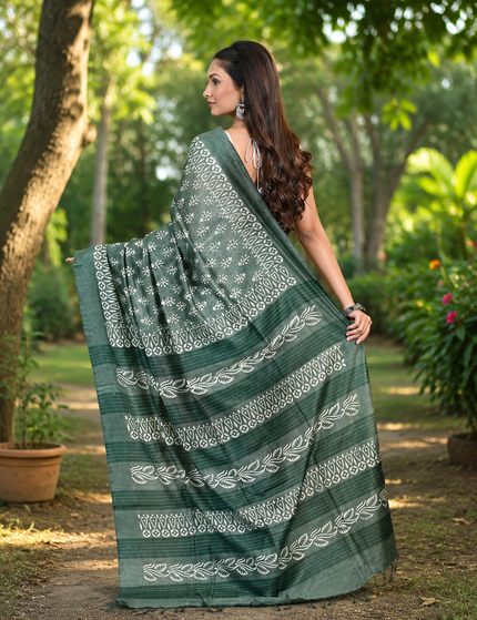 Back view of a woman displaying a green Tussar silk cotton saree with white floral Batik print and decorative border along a garden path.