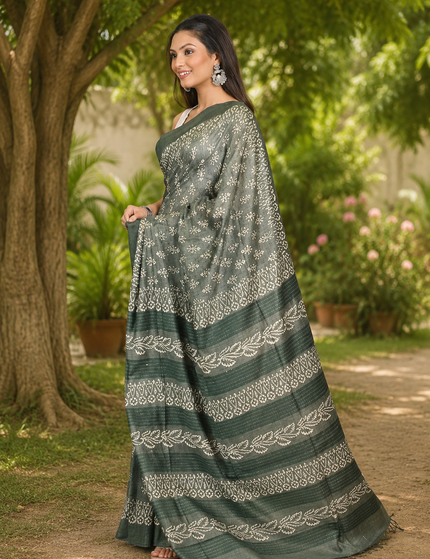 Three-quarter view of a woman wearing a green Tussar silk cotton saree with white floral Batik print and decorative border, standing in a sunlit garden.
