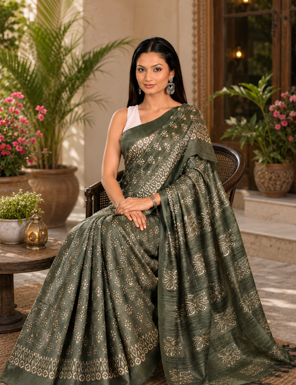 Woman seated in a cane chair wearing a green Tussar silk cotton saree with white floral Batik print and decorative border, with the pallu spread across a patterned rug in a courtyard.