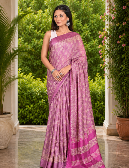 Woman wearing a mauve-lavender Tussar silk cotton saree with delicate white Batik floral print and magenta border, standing in a sunlit garden courtyard.