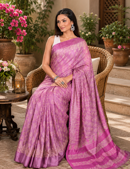 Woman seated in a wicker chair wearing a mauve-lavender Tussar silk cotton saree with delicate white Batik floral print and magenta border, in a sunlit courtyard.