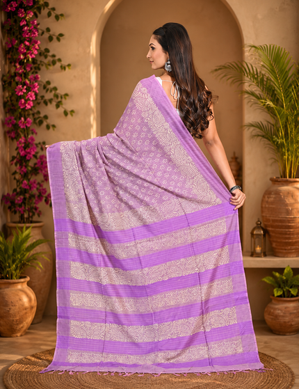 Back view of a woman displaying a pastel lavender Tussar silk cotton saree with delicate white Batik floral print and purple striped border in a sunlit courtyard.