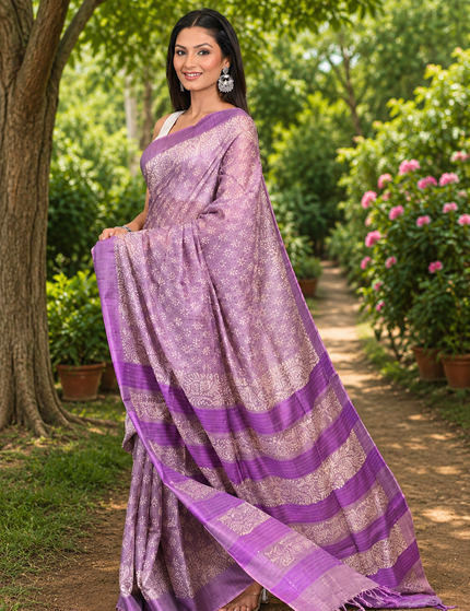 Three-quarter view of a woman wearing a pastel lavender Tussar silk cotton saree with delicate white Batik floral print and purple striped border, standing on a sunlit garden path.