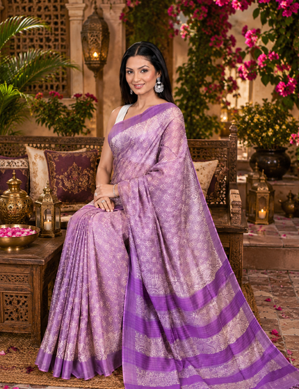 Woman seated on a carved wooden bench wearing a pastel lavender Tussar silk cotton saree with delicate white Batik floral print and purple striped border in a flower-filled courtyard.