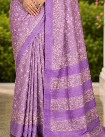 Close-up of a pastel lavender Tussar silk cotton saree showing delicate white Batik floral print, pleats, purple border, patterned bands, and tasseled pallu.