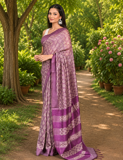 Three-quarter view of a woman wearing a light plum Tussar silk cotton saree with delicate geometric Batik pattern and darker plum striped border, standing on a sunlit garden path.