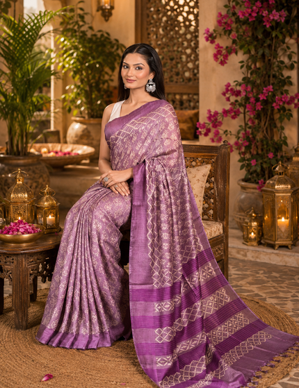 Woman seated in an ornate courtyard chair wearing a light plum Tussar silk cotton saree with delicate geometric Batik pattern and darker plum striped border, with the embellished pallu spread across a woven rug amid lanterns and flowers.