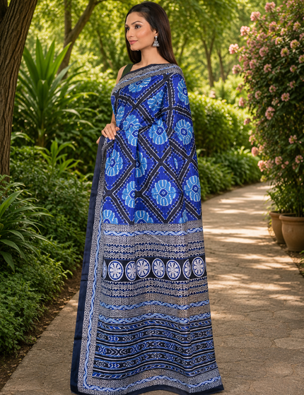 Model wearing a royal blue mercerized Ikkat silk saree, three-quarter front view showing blue floral motifs, diamond patterns, circular details, and black-and-white striped borders on a garden path.