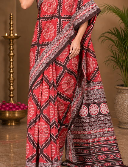 Close-up of a red mercerized Ikkat silk saree showing red floral motifs, geometric diamond patterns, pleats, black-and-white borders, and detailed striped pallu.