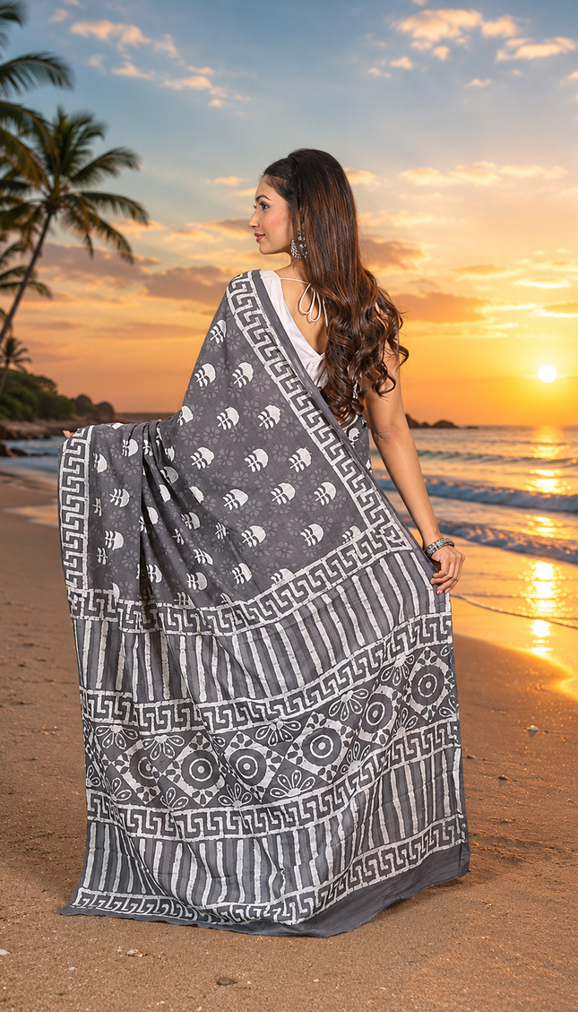 Model showing the back drape of a slate black Jaipur cotton Dabu saree, with white motifs, geometric border, striped bands and circular patterns on a beach at sunset.