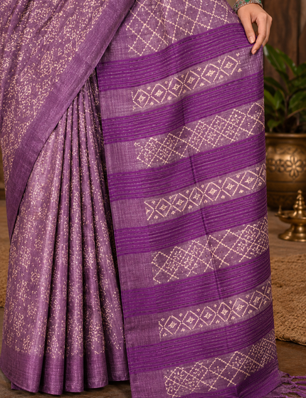 Close-up of a light plum Tussar silk cotton saree showing delicate geometric Batik motifs, pleats, darker plum striped border, woven texture, and tasseled pallu in a warm courtyard setting.