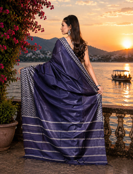 Back view of a woman displaying a navy blue cotton Fishbone saree with silver chevron border and striped pallu beside a lake at sunset.