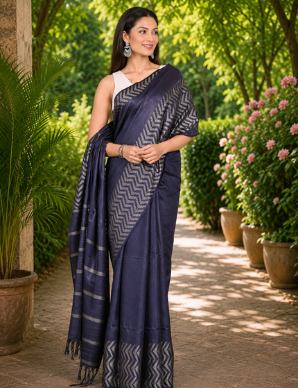 Three-quarter view of a woman wearing a navy blue cotton Fishbone saree with silver chevron border and striped pallu, standing on a sunlit garden path.