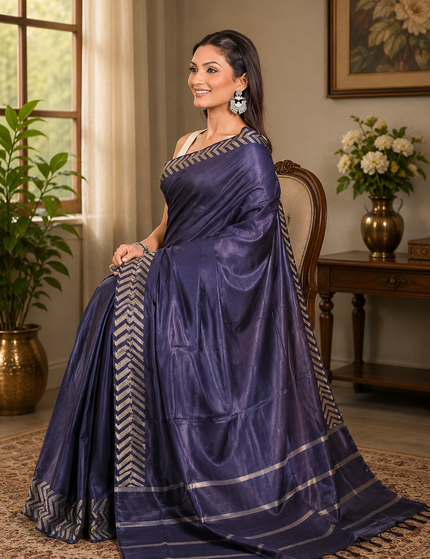 Woman seated on an upholstered chair wearing a navy blue cotton Fishbone saree with silver chevron border and striped pallu, with the saree spread across a patterned rug in an elegant room.