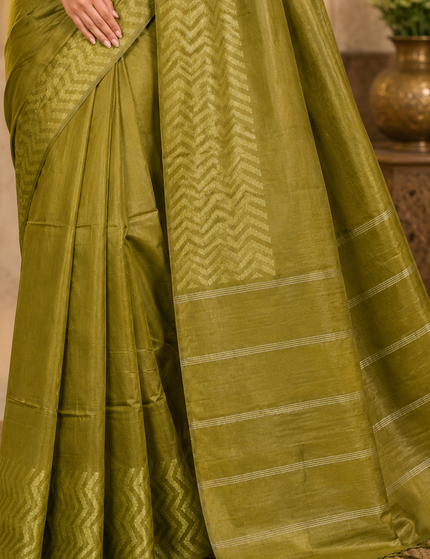 Close-up of an olive green cotton Fishbone saree showing the zigzag herringbone border, fine striped bands, pleats, and tasseled pallu indoors.