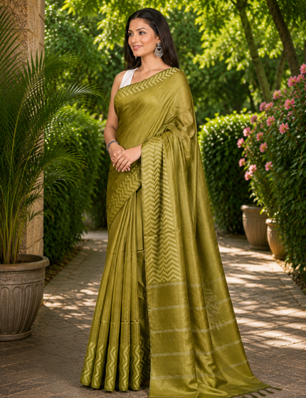 Three-quarter view of a woman wearing an olive green cotton Fishbone saree with a zigzag herringbone border and striped pallu, standing on a sunlit garden path.
