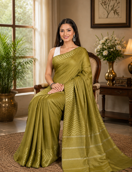 Woman seated in an armchair wearing an olive green cotton Fishbone saree with a zigzag herringbone border and striped pallu in a softly lit room.