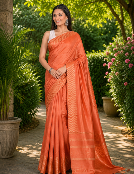 Woman wearing a coral orange cotton Fishbone saree with a zigzag herringbone border and striped pallu, standing in a sunlit garden.