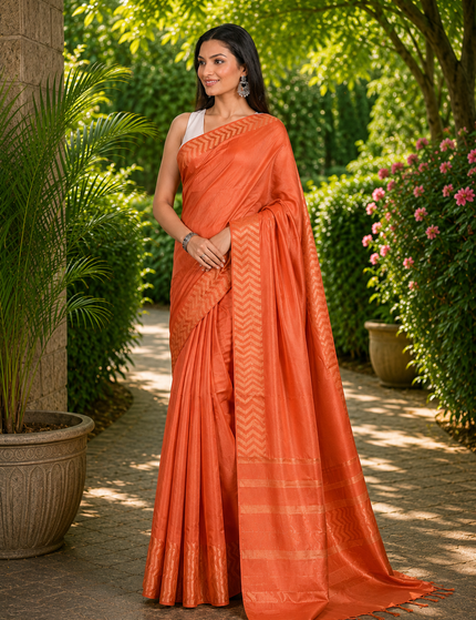 Three-quarter view of a woman wearing a coral orange cotton Fishbone saree with a zigzag herringbone border and striped pallu, standing on a sunlit garden path.