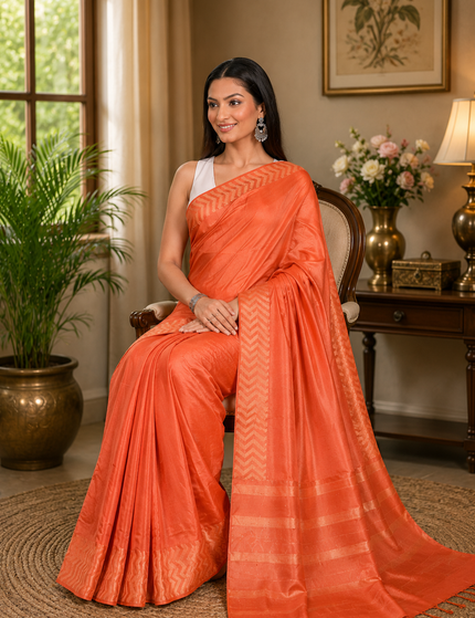 Woman seated in an armchair wearing a coral orange cotton Fishbone saree with a zigzag herringbone border and striped pallu in a softly lit room.
